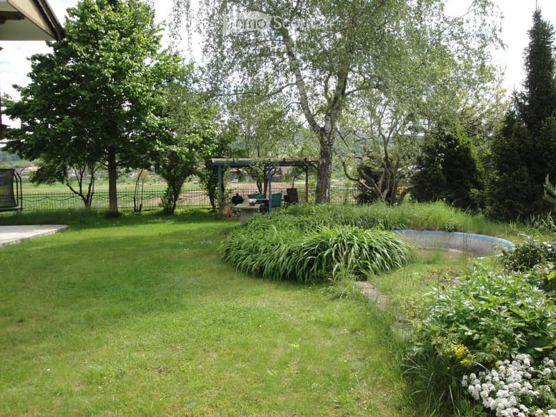 Well-maintained garden with a lawn, various plants, a small pond, a covered seating area, and a fence surrounding the property.