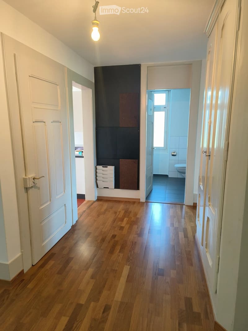 A well-lit hallway with wooden flooring, white walls, and several doors leading to different rooms, including a visible bathroom with a toilet and a small storage unit.