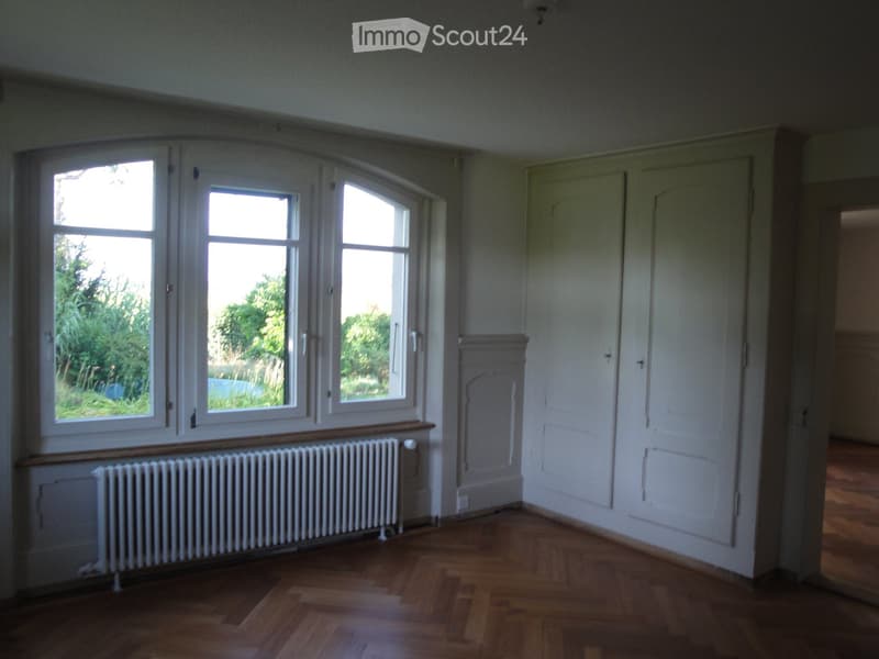 Empty room with wooden flooring, white walls, large windows with white frames, radiator, and two white doors.