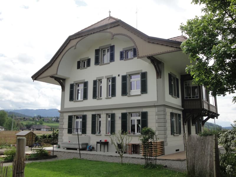 Two-story house with white walls, dark shutters, and a balcony on the second floor. The house features a well-maintained garden with a small patio area, outdoor furniture, and a wooden fence.
