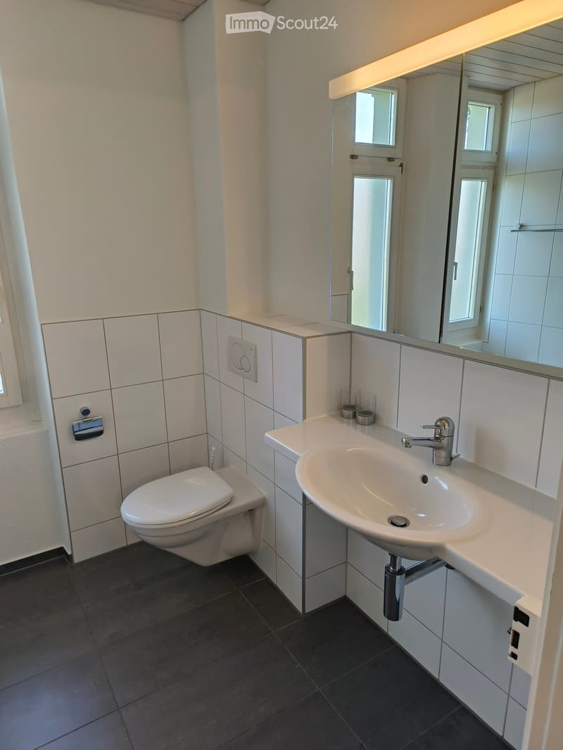 Modern bathroom with white tiles, a toilet, a sink with a modern faucet, a large mirror, and two small cups on the counter.