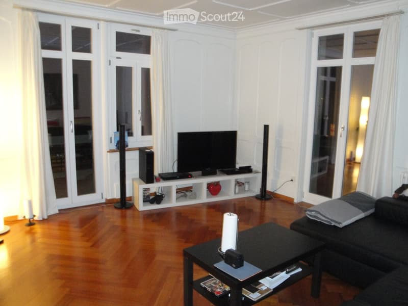 Modern living room with hardwood flooring, large windows, a black sofa, a coffee table, a TV stand with a flat-screen TV, and speakers.