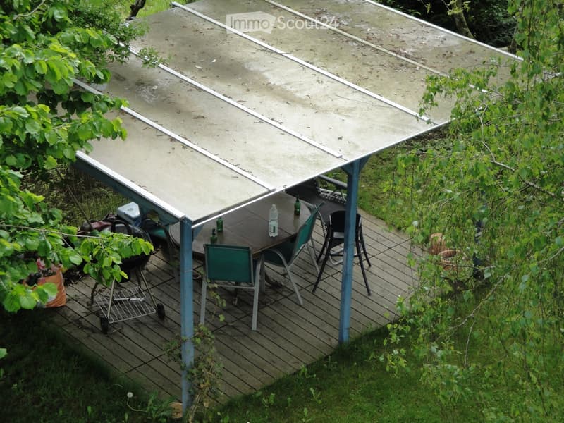 A wooden deck with a covered seating area, including a table, chairs, and a grill, surrounded by greenery and a grassy area.