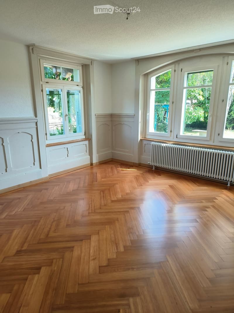 Empty room with polished wooden herringbone flooring, large windows with white frames, radiator, and decorative wall paneling.