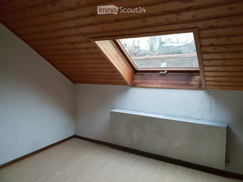 Empty attic room with wooden slatted ceiling, skylight window, white walls, and a radiator.