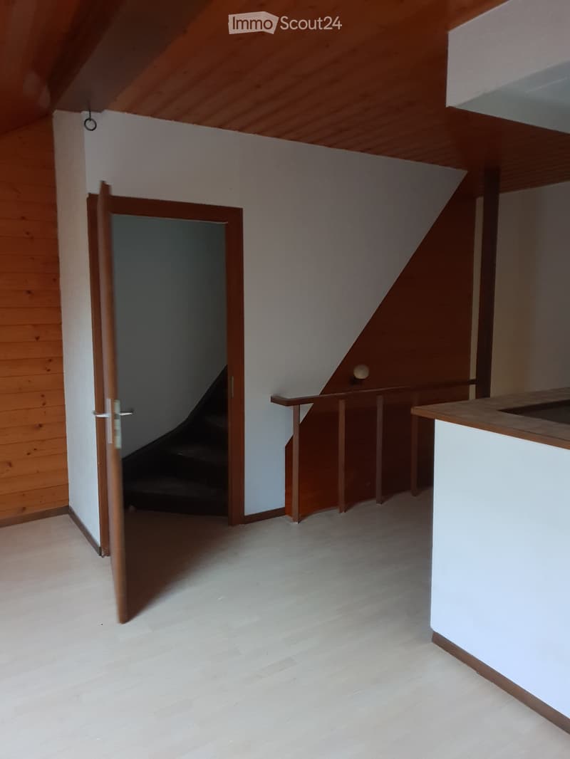 Empty room with wooden floor, white walls, and a wooden door leading to a staircase. The ceiling is slanted with wooden beams.