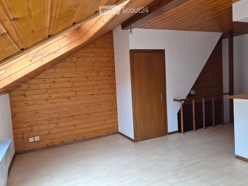 Empty room with wooden walls, slanted wooden ceiling, and a wooden door. The floor is light-colored wood, and there is a small staircase leading upwards.