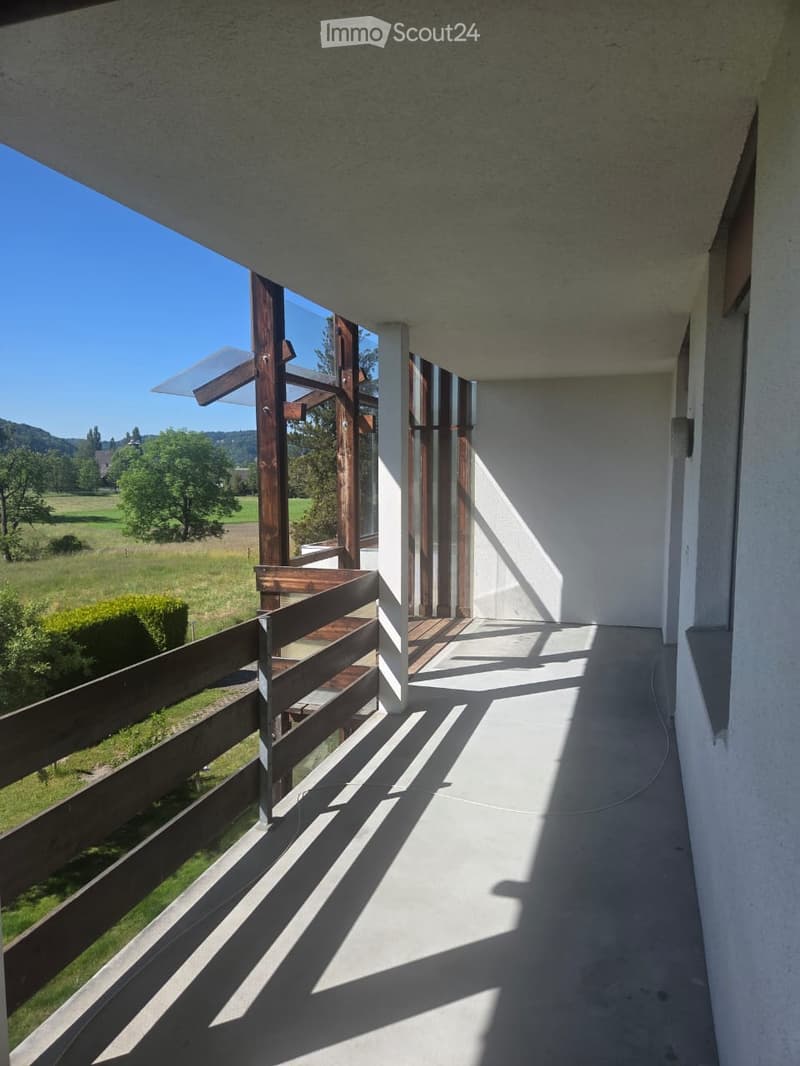 Balcony with wooden railing, concrete floor, and scenic view of a green field and trees.