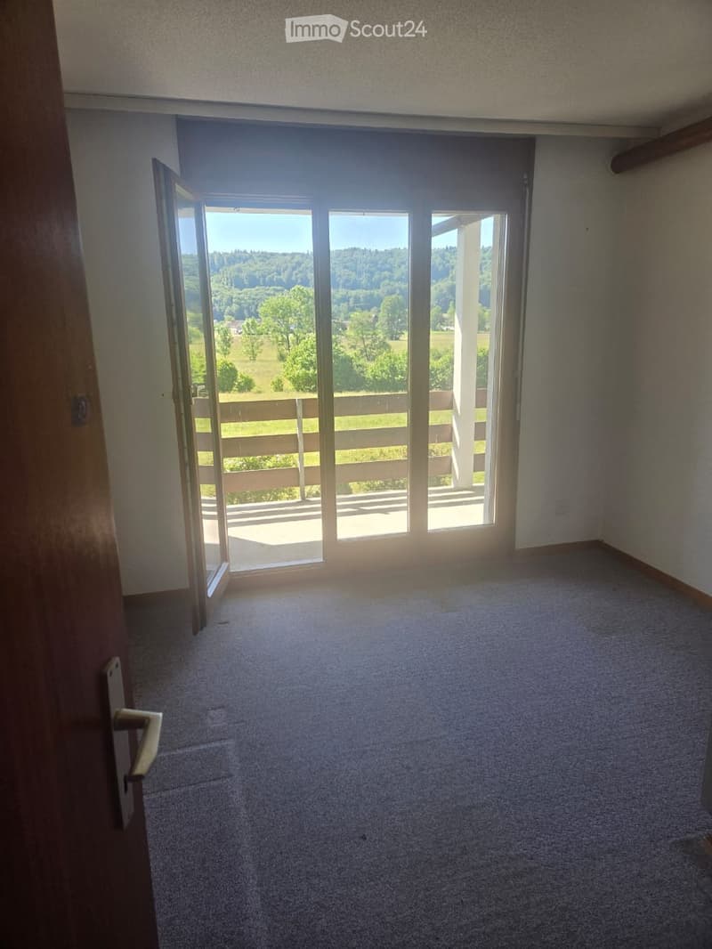 Empty room with carpeted floor, large sliding glass door opening to a balcony with a scenic view of a green landscape.