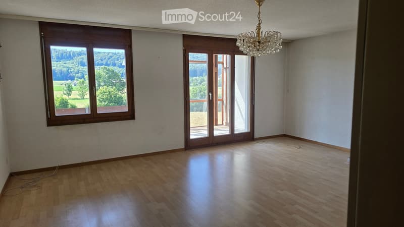 Empty room with wooden flooring, large windows, and a chandelier hanging from the ceiling. The room has white walls and a sliding glass door leading to a balcony.