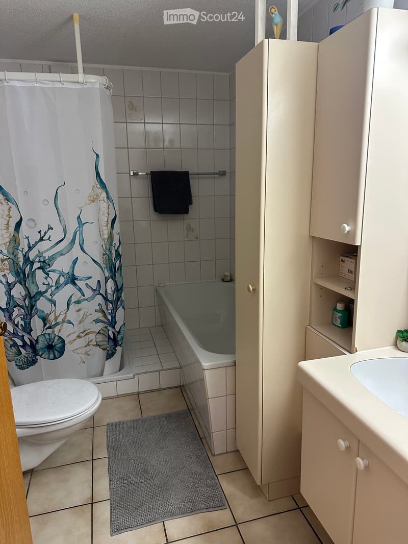 Bathroom with a toilet, bathtub, sink, and a cabinet. Features a shower curtain with a sea-themed design, a towel on the rack, and a small rug on the floor.