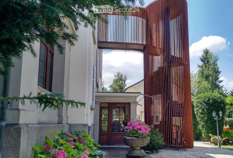 A modern building with a wooden lattice structure and a balcony, featuring a sign reading "Sonnenheim" above the entrance, surrounded by greenery and flowers.