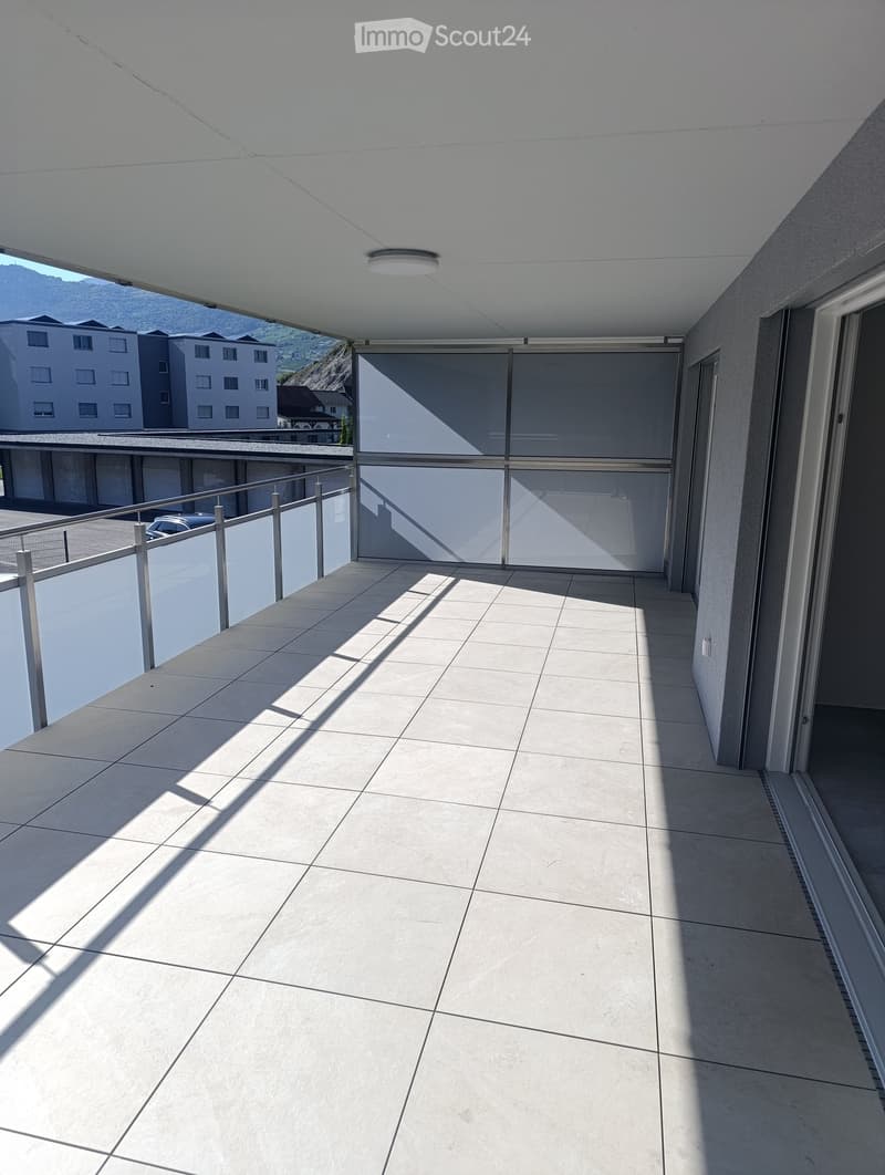 Modern balcony with tiled floor, metal railing, and large glass sliding doors leading to the interior. The balcony has a clean, minimalist design with a view of surrounding buildings and mountains.