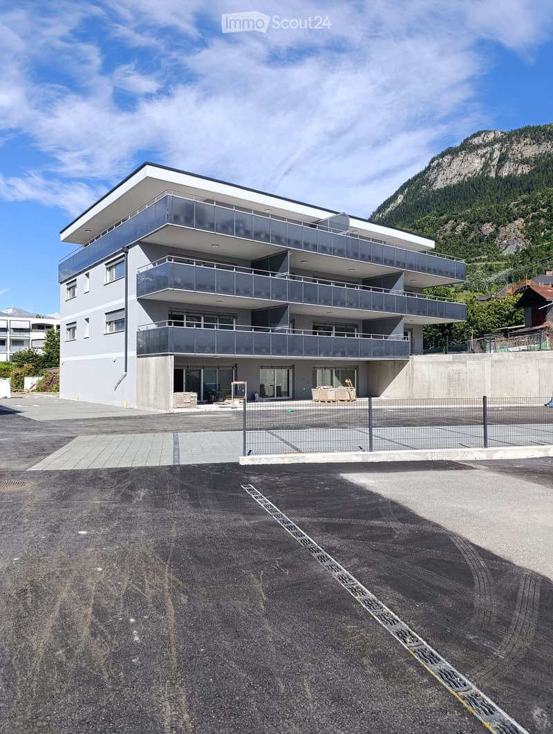 Modern three-story building with balconies, large windows, and a contemporary design, situated in a parking lot with a mountain backdrop.