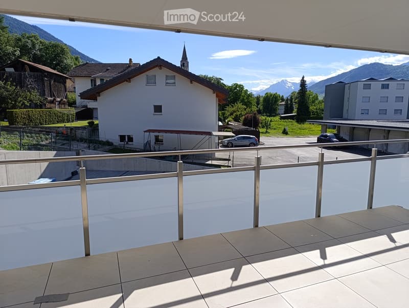 Balcony with metal railing overlooking a scenic town with houses, mountains, and a church spire in the background.