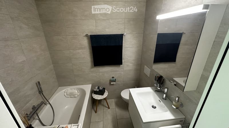 Modern bathroom with beige tiled walls and floor, featuring a bathtub, sink, toilet, and a small wooden stool holding a decorative item.