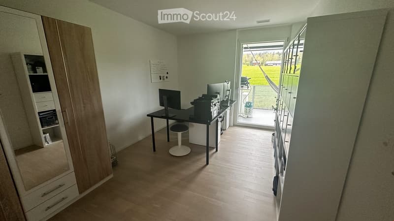 A modern, well-lit room with a desk, chair, computer, printer, and a tall wardrobe with shelves. The room has light wooden flooring and large windows offering a view of a green field.