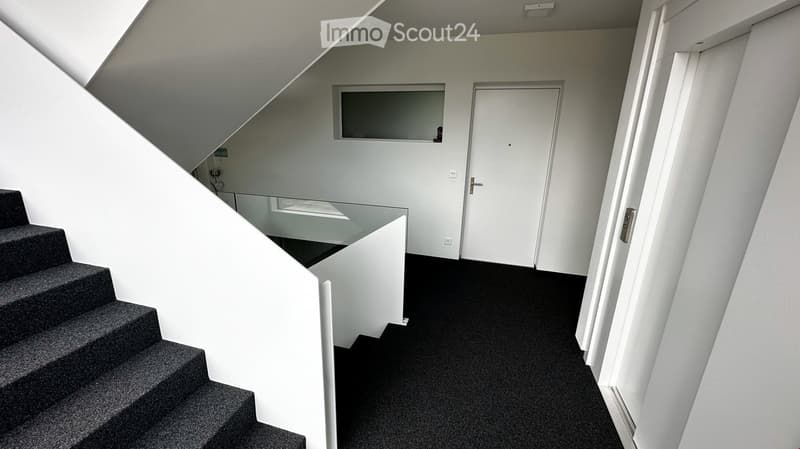 Modern hallway with carpeted stairs, white walls, and a glass railing leading to an open area. A closed door is visible at the end of the hallway.