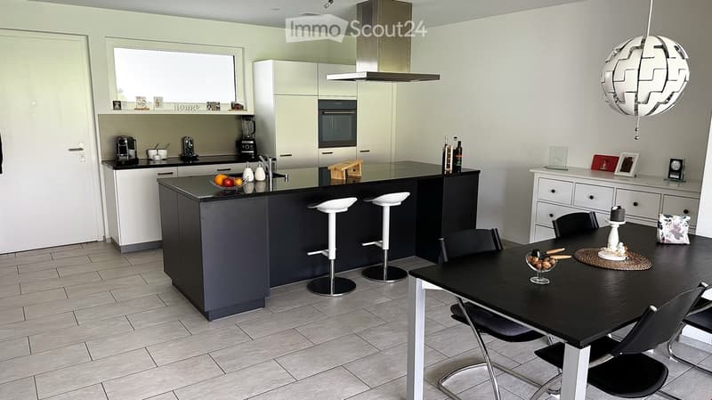 Modern kitchen with white cabinets, black island, and integrated appliances. Features a dining area with a black table and chairs, a hanging lamp, and various kitchen items on the counter.