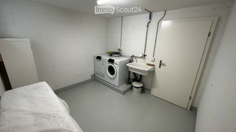 Small room with a bed, washing machine, sink, and toilet. The walls are white brick, and the floor is concrete.