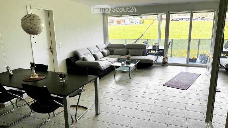 Modern living and dining area with a grey sectional sofa, glass coffee table, black dining table with chairs, and a hanging spherical lamp. Large sliding glass doors open to a view of a green field.