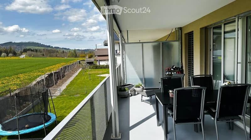 Balcony with outdoor furniture, including a table, chairs, and potted plants, overlooking a scenic countryside view.