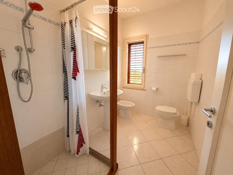 Modern bathroom with white tiles, a toilet, a sink, a shower with a curtain, and a window with blinds.