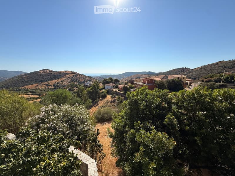 A scenic view of a rural area with rolling hills, scattered houses, and lush vegetation, including trees and shrubs, under a clear blue sky.