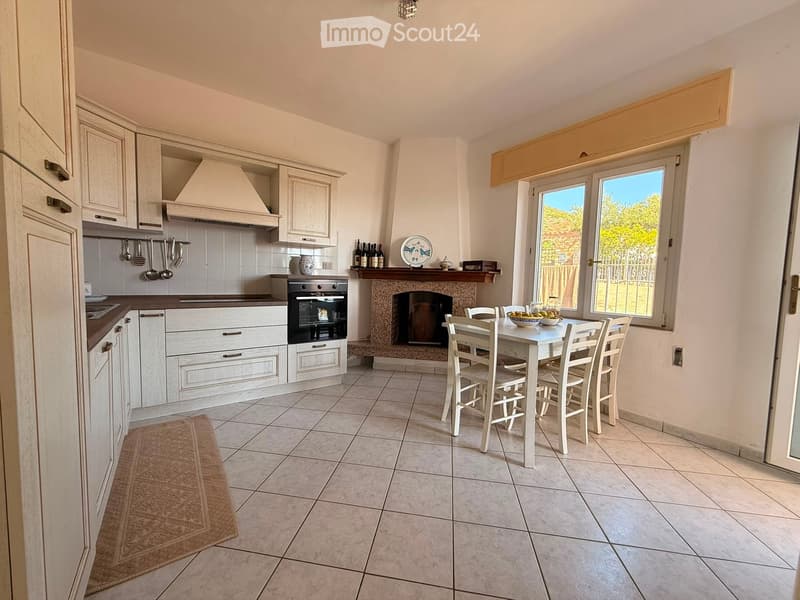 Bright kitchen with white cabinets, a central island, a built-in oven, a fireplace, and a dining area with a table and chairs. The space features tiled flooring and large windows offering a view of the outdoors.