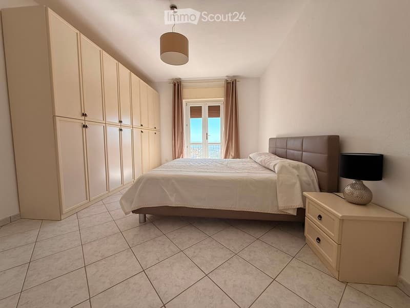 Modern bedroom with a large bed, nightstand, lamp, and built-in wardrobe. The room features tiled flooring and large windows with curtains.