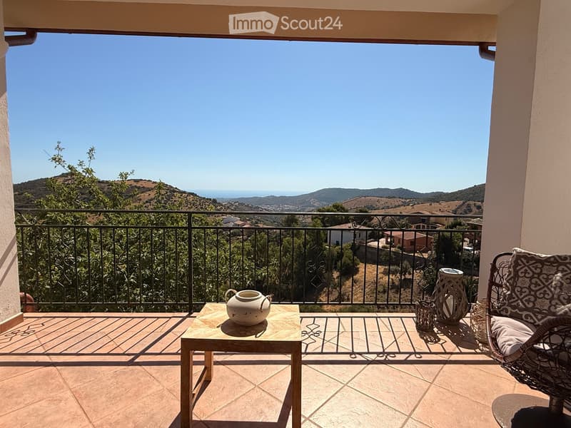 Balcony with a scenic view of hills and ocean, featuring a small wooden table, a wicker chair, and decorative vases.