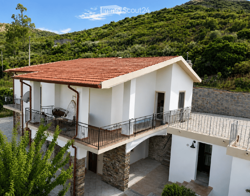 Two-story white house with red-tiled roof, stone accents, and balconies overlooking a green, hilly landscape.
