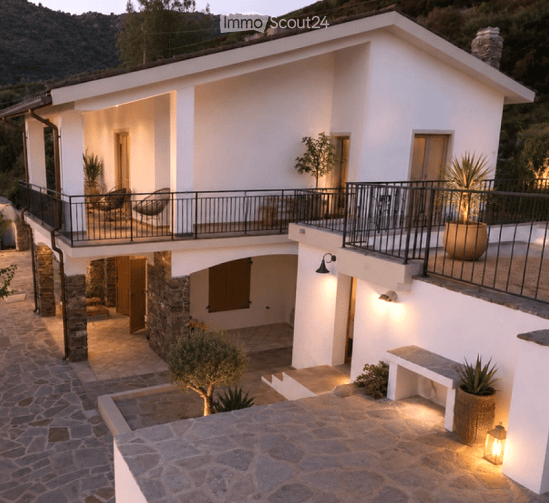 Two-story white house with a balcony, stone base, and outdoor seating area.