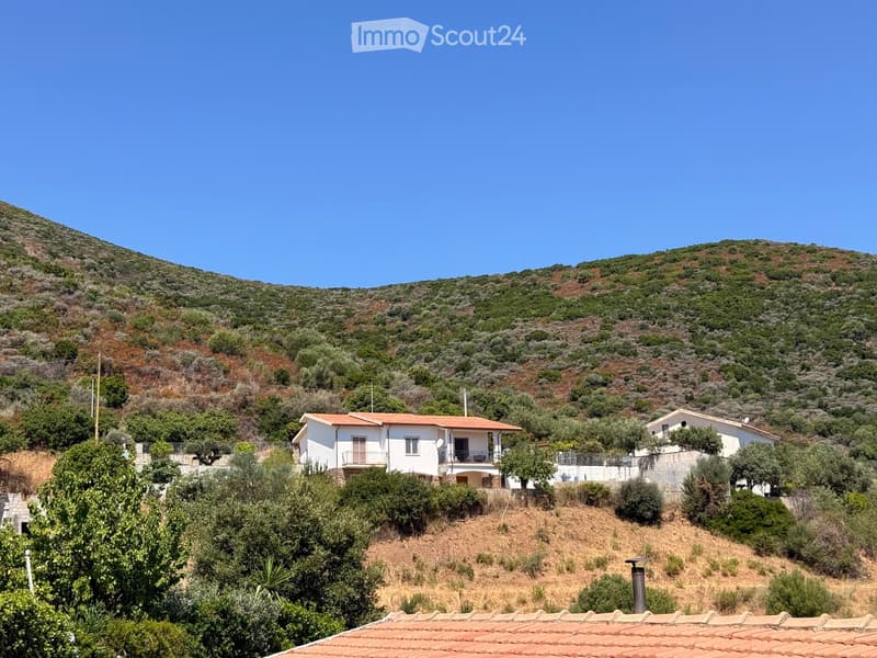 A white two-story house with a red-tiled roof, situated on a hillside with surrounding greenery and distant hills.