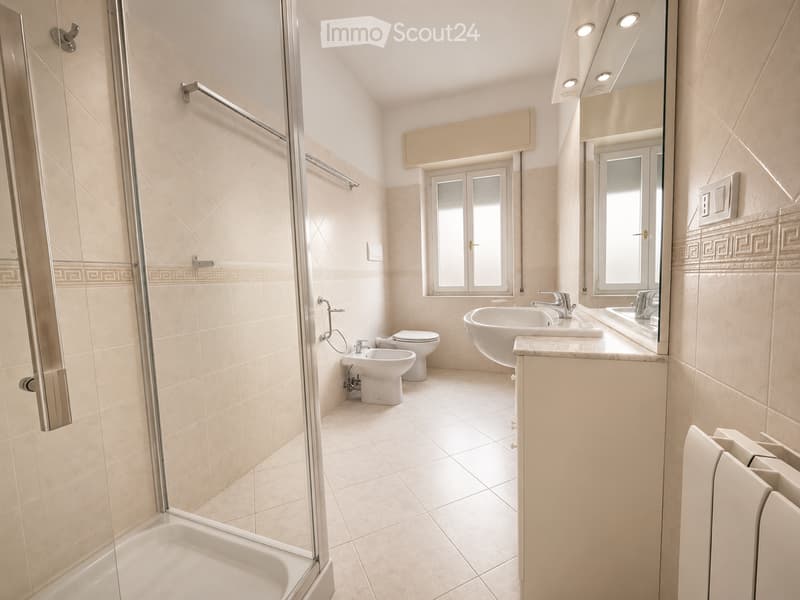 Modern bathroom with beige tiles, a glass-enclosed shower, a bathtub, a toilet, a bidet, and a double sink with a large mirror above it.