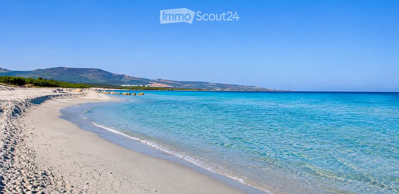 A serene beach with fine white sand, clear turquoise water, and distant hills under a bright blue sky.