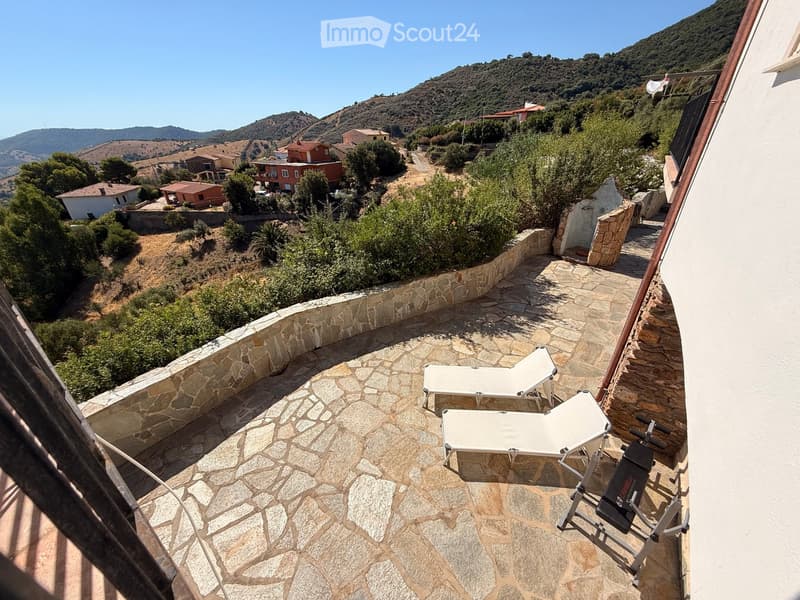 Balcony with stone flooring, two sun loungers, and a fitness machine, overlooking a scenic hillside with houses and greenery.