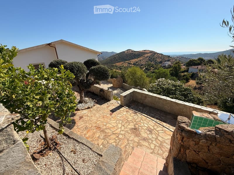 A stone-paved outdoor area with a small tree, stone walls, and a scenic view of hills and the sea in the background.