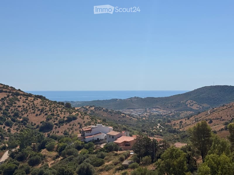 A panoramic view of a residential area nestled in a hilly landscape, with several houses and a clear view of the sea in the distance.
