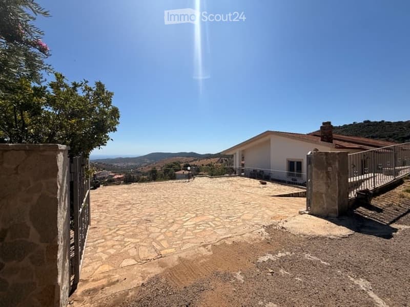 A spacious outdoor area with a stone-paved courtyard, a white house in the background, and a scenic mountain view under a clear blue sky.