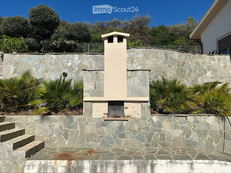 Stone wall with a small chimney structure, surrounded by palm trees and greenery, with steps leading up to the wall.