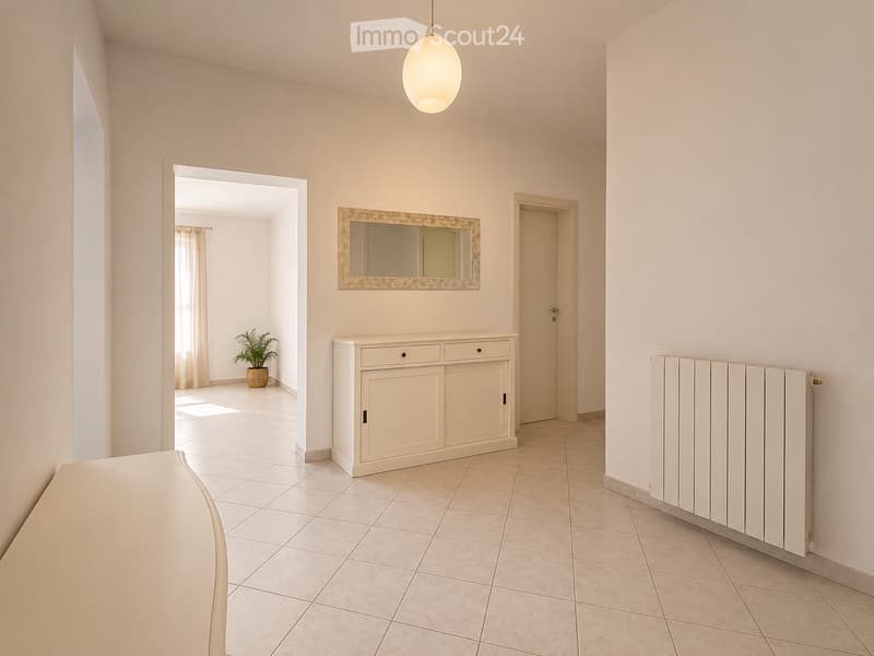 Bright hallway with tiled flooring, white walls, a hanging light fixture, a small cabinet, a mirror, a radiator, and a potted plant visible in the adjacent room.
