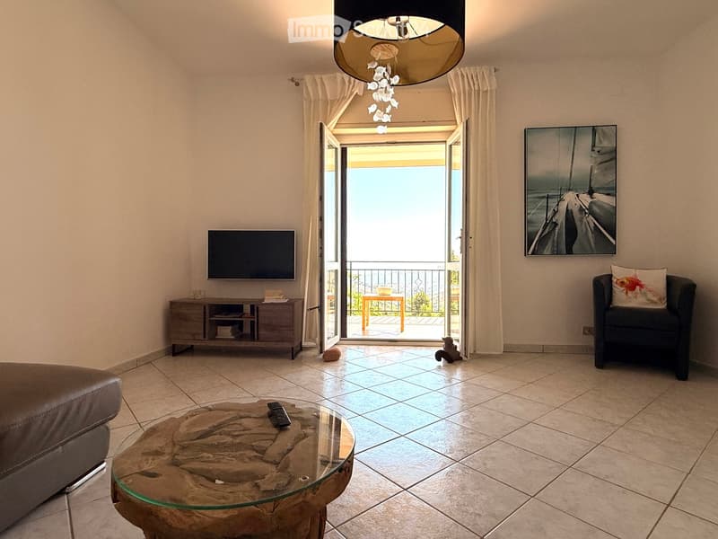 Modern living room with tiled floor, large windows, balcony access, sofa, coffee table, TV, and artwork.