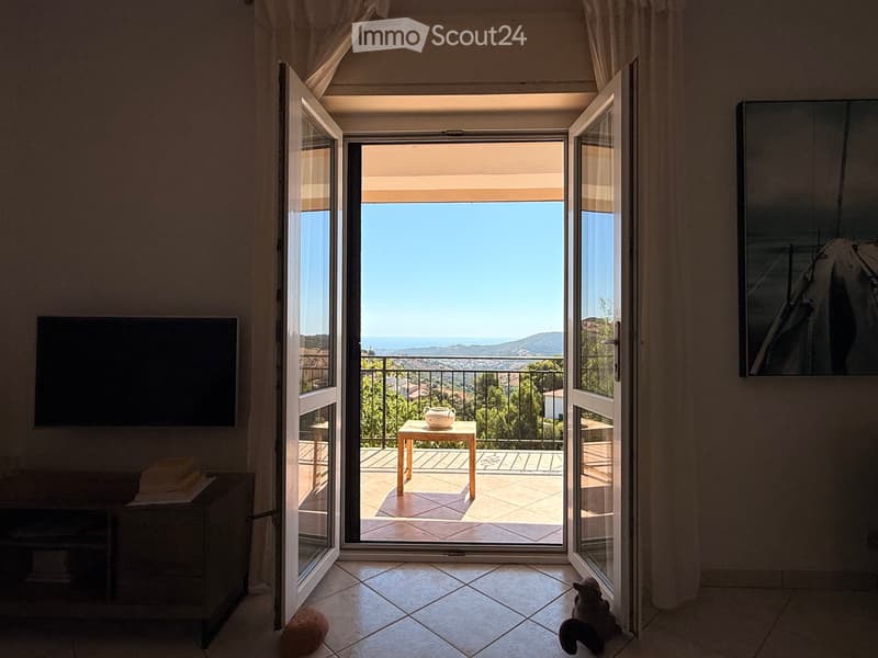Bright room with large sliding glass doors opening to a balcony with a scenic view. Inside, a small table with a teapot sits on the balcony, and a screen is mounted on the wall. A small pet is visible near the floor.