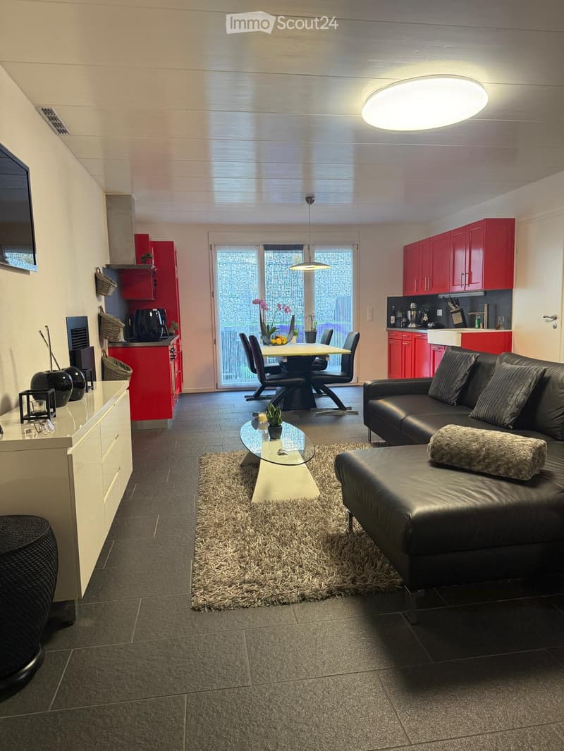 Modern living and dining area with a black leather sofa, glass coffee table, ottoman, and a dining table with chairs. The kitchen features red cabinets and stainless steel appliances. The space is well-lit with large windows and a circular ceiling light.