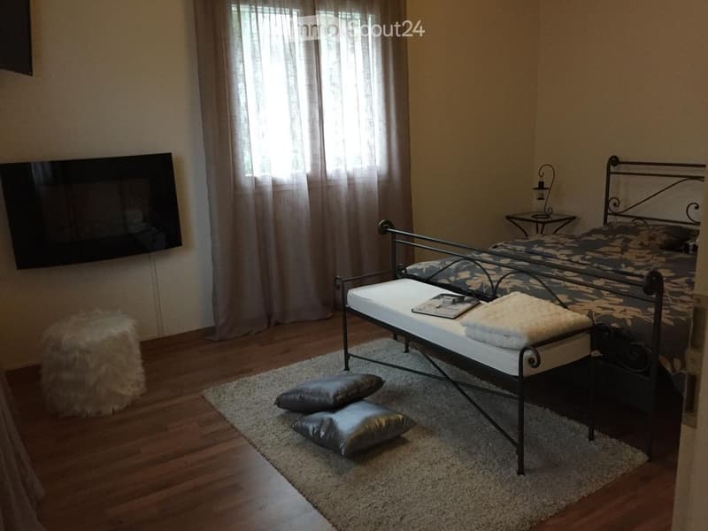 Cozy bedroom with wooden flooring, a metal-framed bed, a bench at the foot of the bed, a nightstand with a table lamp, and a large mirror on the wall.