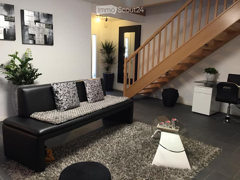 Modern living room with a black leather sofa, patterned cushions, a glass coffee table, a grey shag rug, a black ottoman, a white side table, a black office chair, and a staircase leading to another level.