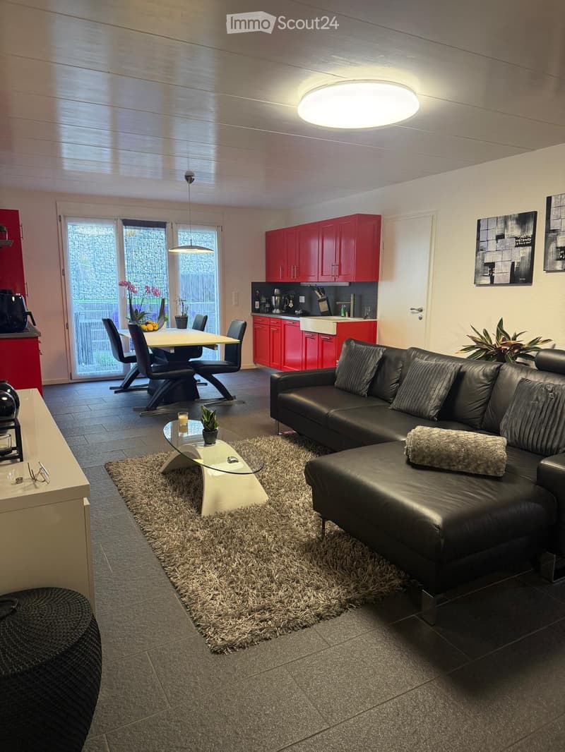 Modern living room with a black leather sofa, glass coffee table, dining area with chairs and table, red cabinets, and a large window with a view of the garden.