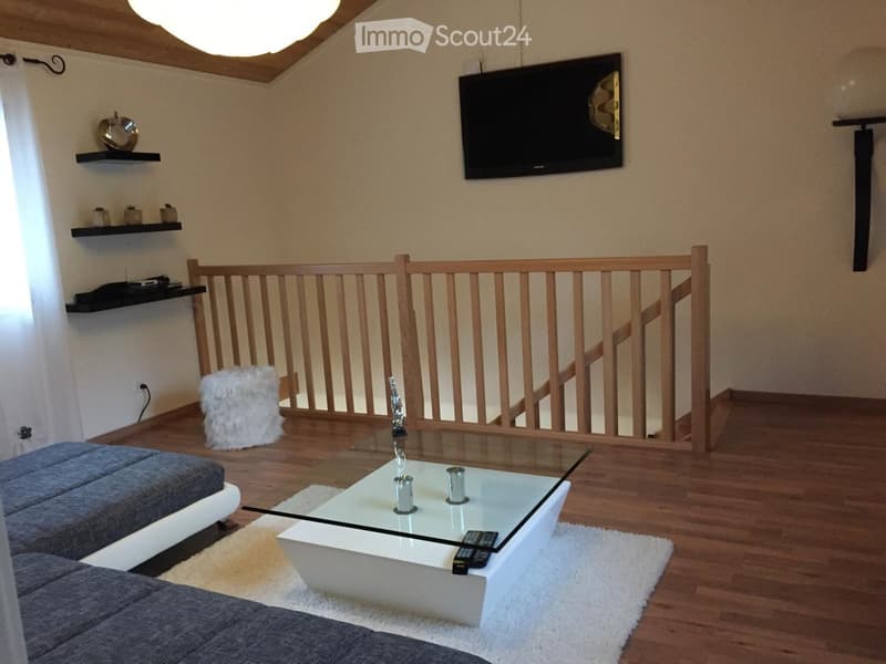 Modern living room with wooden flooring, a grey sofa, a white coffee table with remote controls, shelves with decorative items, and a mounted television. A staircase with wooden railings is visible in the background.