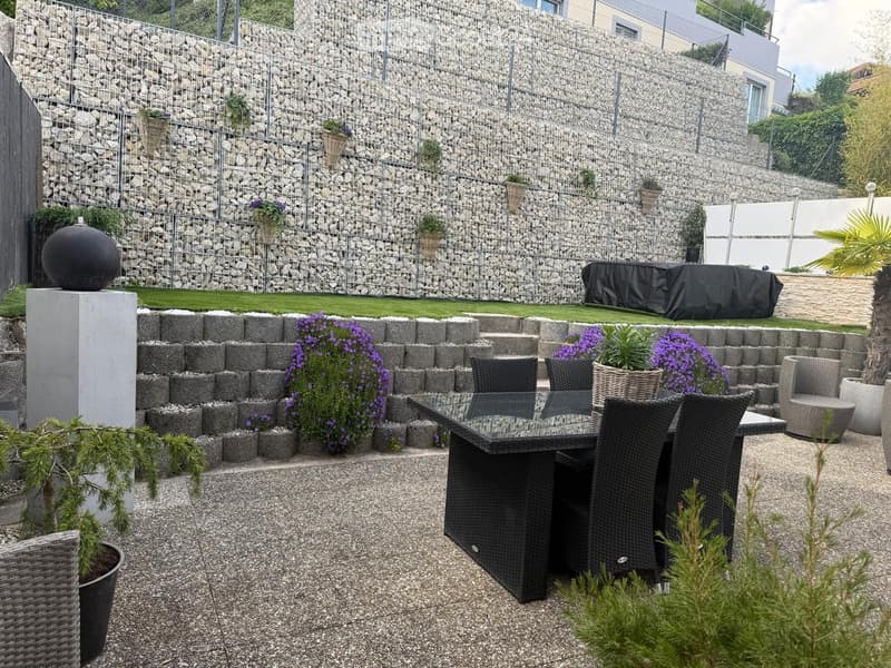 Well-maintained garden with a stone wall, gravel pathway, wicker furniture, and potted plants.